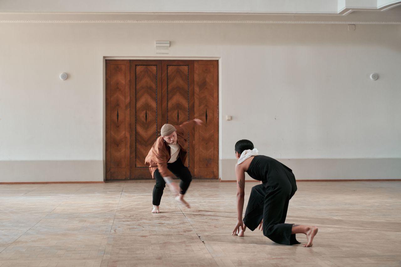 Two dancers performing contemporary dance in a minimalist indoor setting.